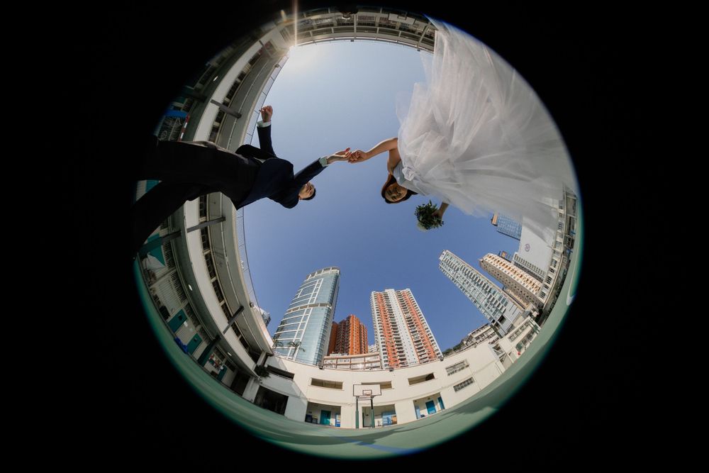 Creative fisheye lens wedding photo of couple holding hands against a blue sky with city skyscrapers.