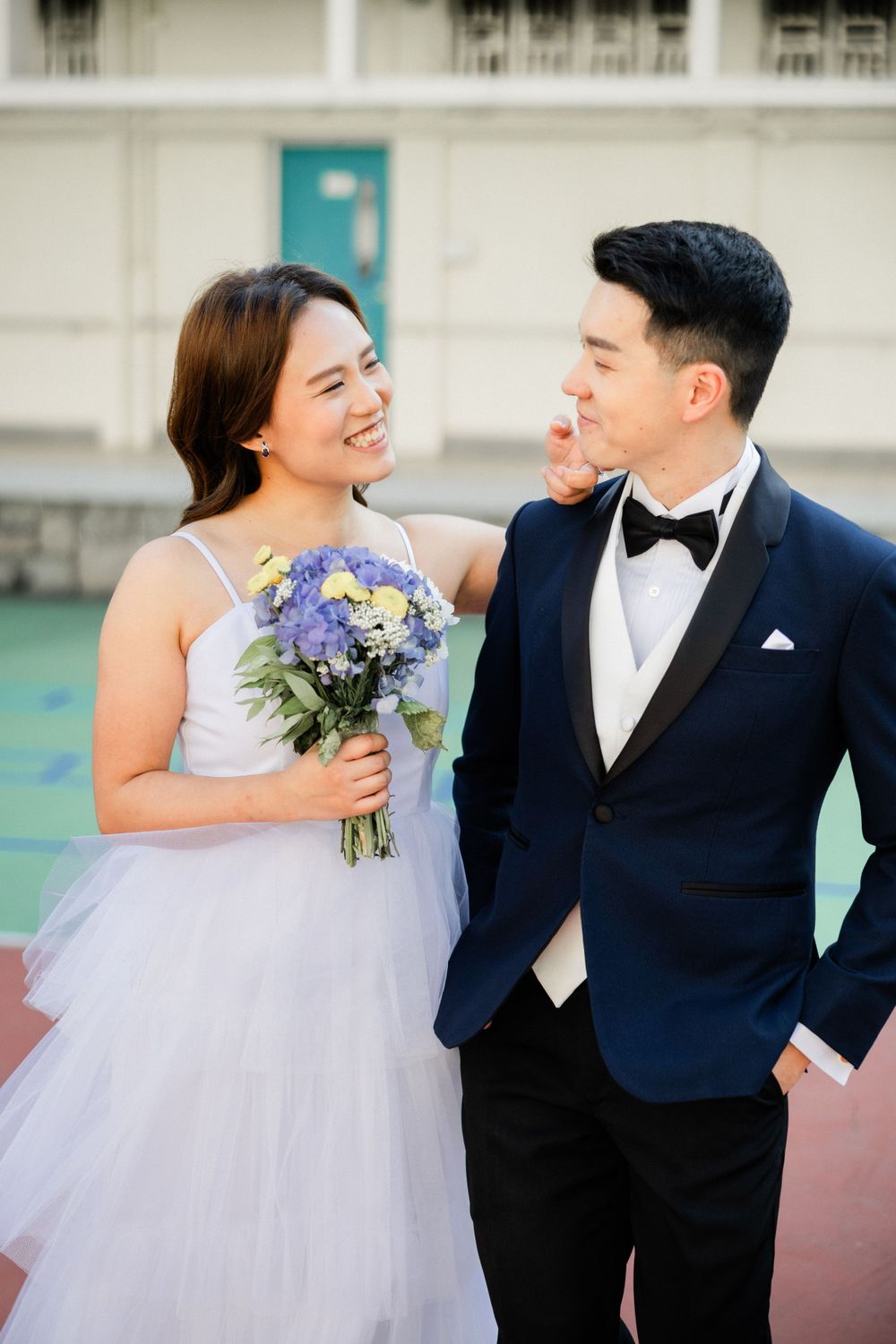 Smiling Asian bride in white dress holding purple bouquet gazes at groom in navy tuxedo on outdoor court.