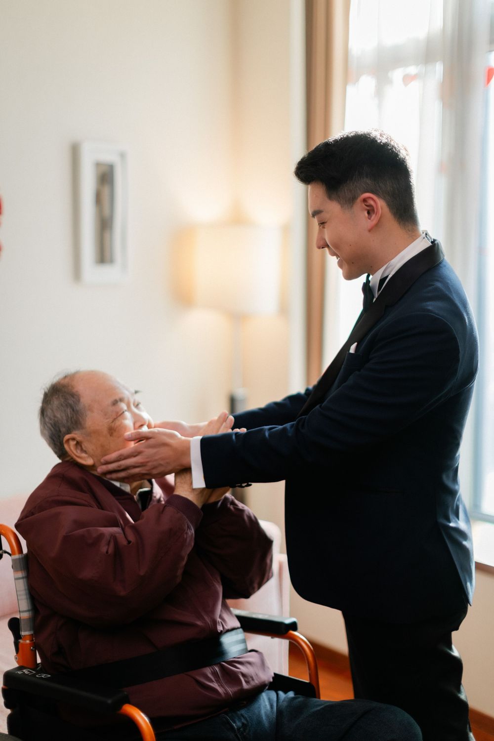 Smiling groom in navy tuxedo tenderly holds the face of an elderly man seated in a wheelchair indoors.