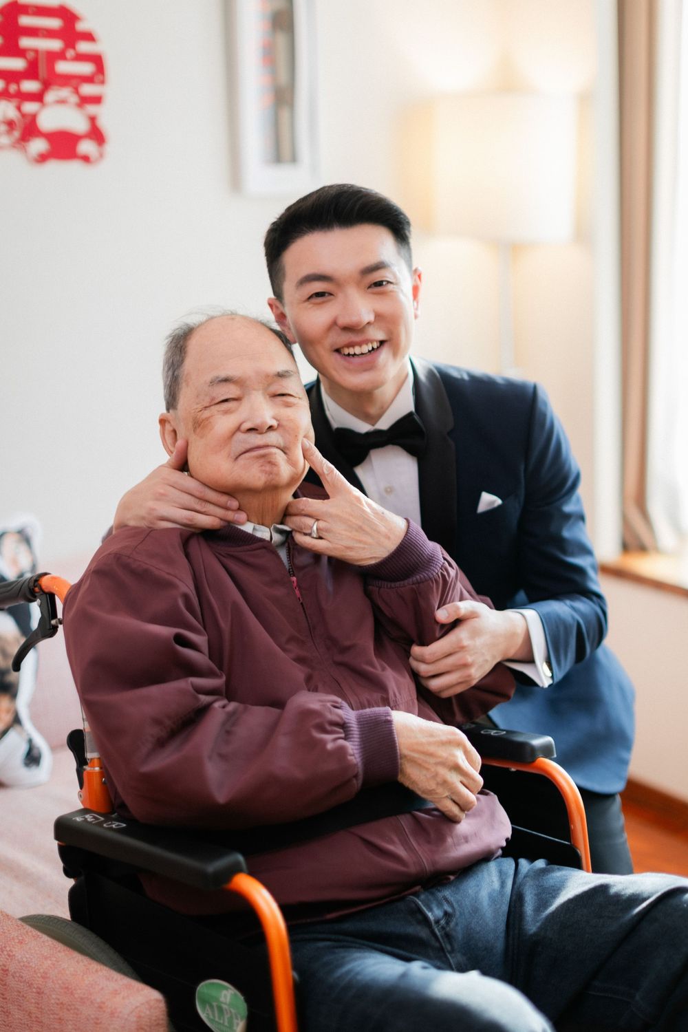 Joyful groom in navy tuxedo embraces smiling elderly man in wheelchair during wedding day family visit.