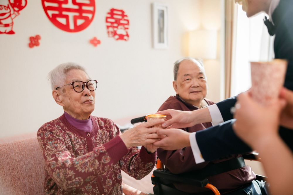 Elderly couple receiving tea cups during a traditional Chinese wedding tea ceremony with red double happiness decor.