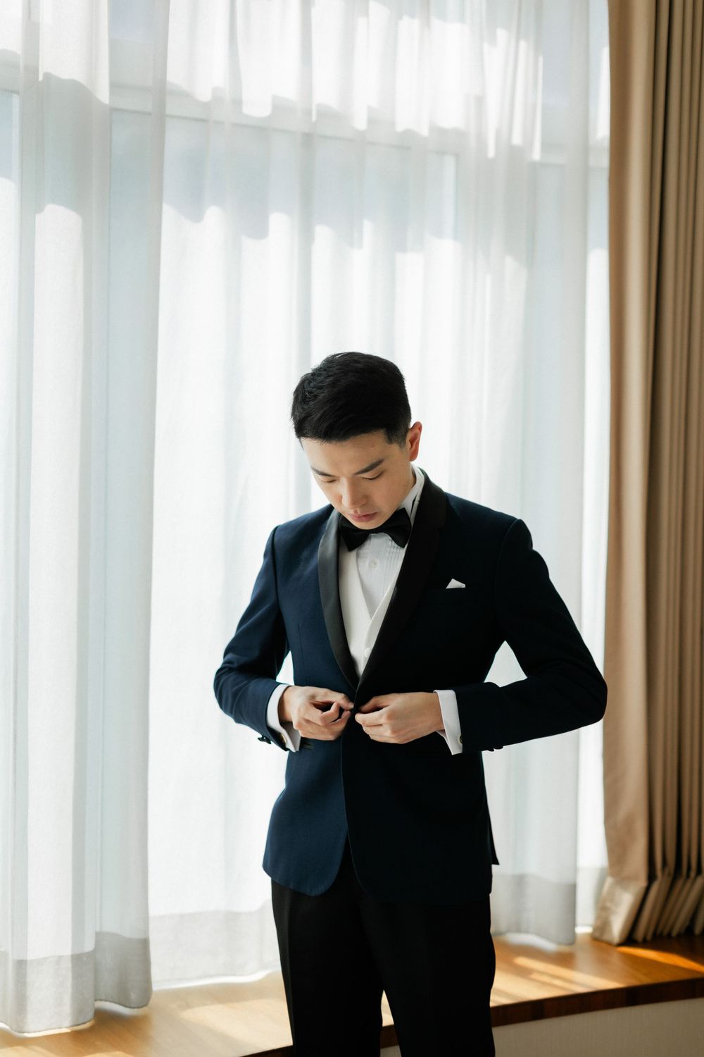 Handsome Asian groom in navy tuxedo buttoning his jacket while standing by sheer curtains.