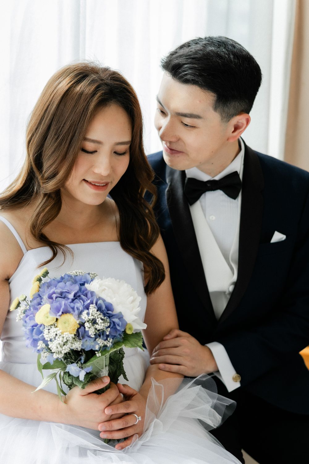 Romantic bride holding blue hydrangea bouquet while groom in tuxedo gazes at her lovingly.