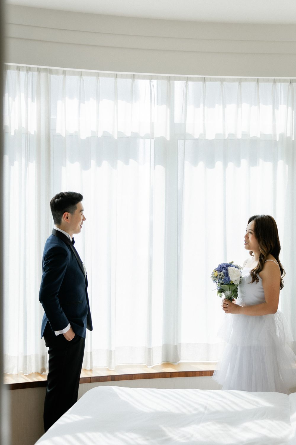Bride and groom share a tender first look moment by a sunlit window, she holds a blue and white bouquet.