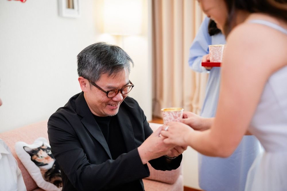 An older man in a black jacket places a gold bracelet on a woman's wrist during a traditional Chinese wedding tea ceremony.