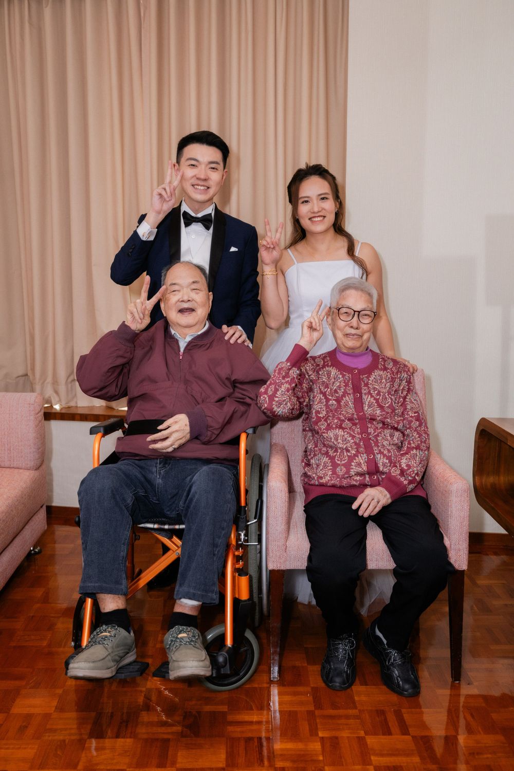 A newlywed couple in wedding attire poses with two elderly grandparents, one in a wheelchair, making peace signs.