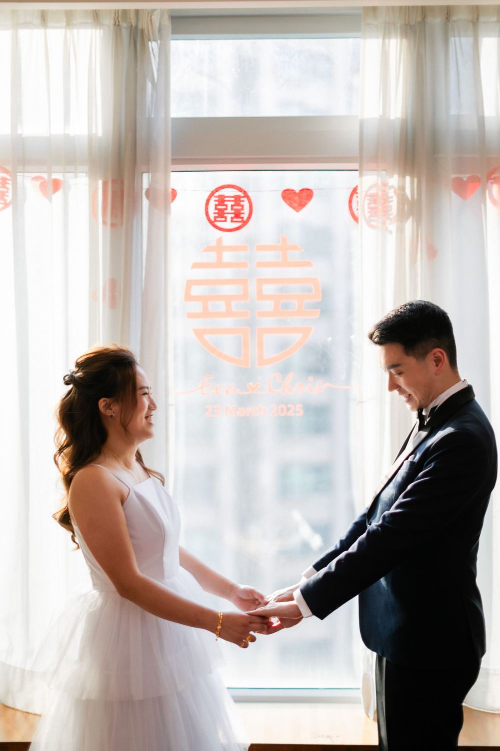 A bride and groom hold hands facing each other before a window decorated with a red Chinese double happiness symbol.