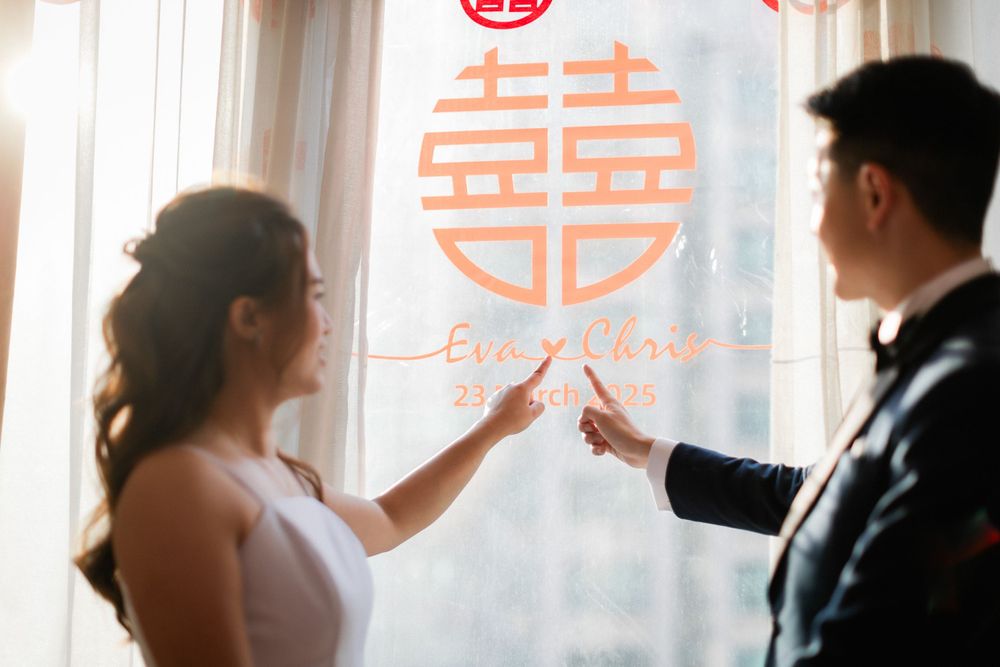 A bride and groom point to a personalized red Chinese double happiness wedding window decoration with their names.