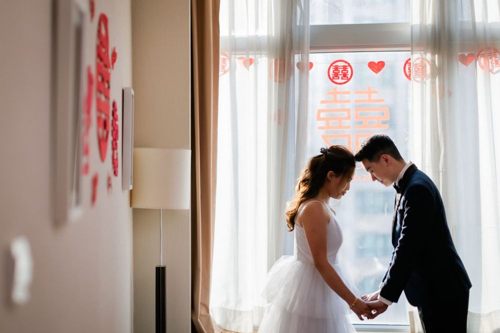 A bride and groom share an intimate forehead-touching moment by a window adorned with red Chinese wedding decorations.