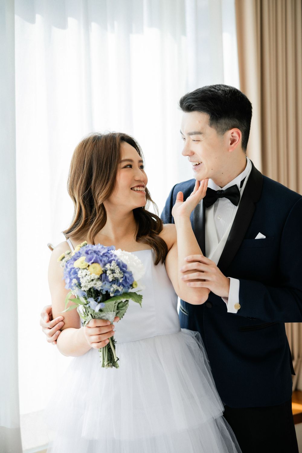 Smiling Asian wedding couple embrace indoors, bride holds blue floral bouquet while groom wears navy tuxedo.