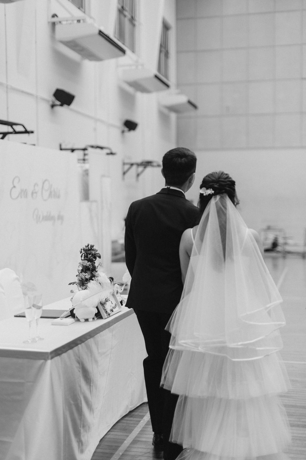 Black and white photo of bride and groom walking toward wedding cake table during ceremony.