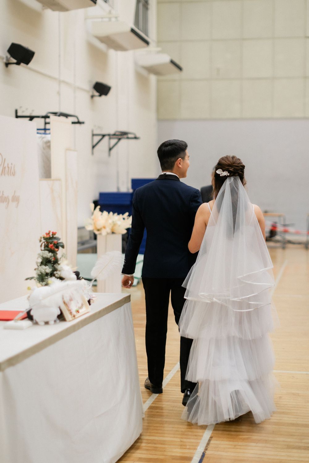 Bride in tiered veil and groom in navy suit walk together past decorated wedding table.