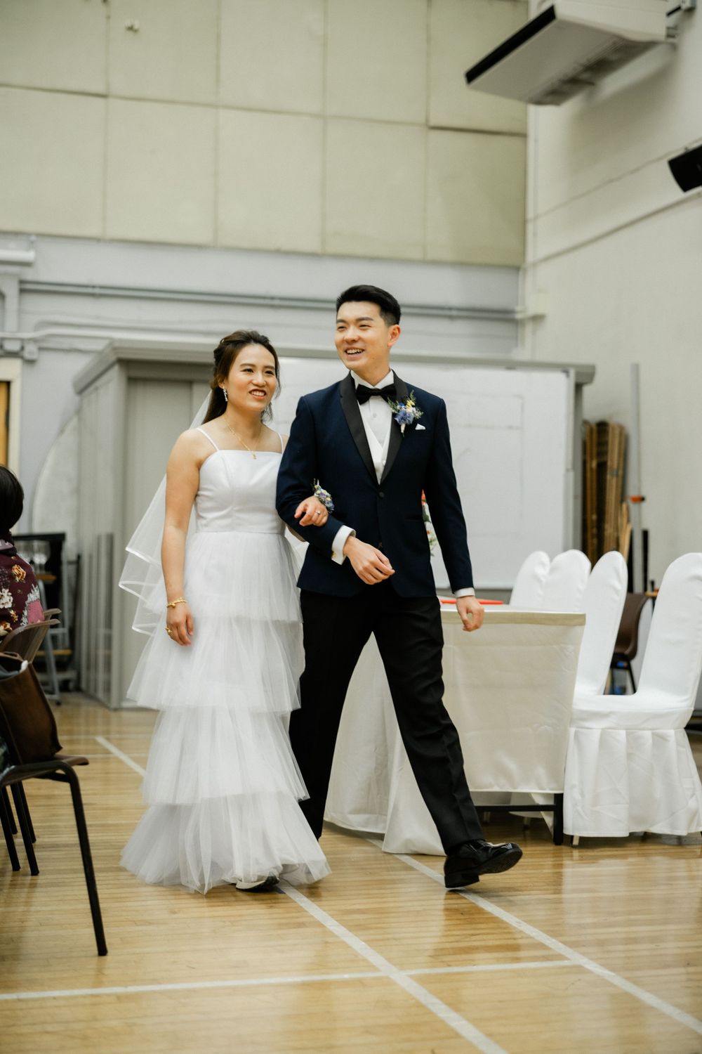 Smiling bride and groom in navy tuxedo walk hand in hand through wedding reception hall.