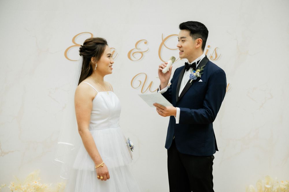 Groom in navy tuxedo reads wedding vows to smiling bride at altar with floral backdrop.