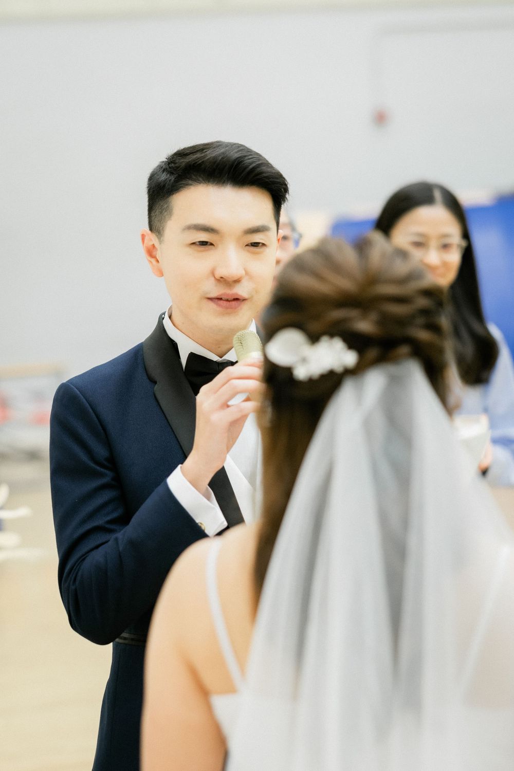 Groom in navy suit speaks vows to bride with floral hair accessories during ceremony.