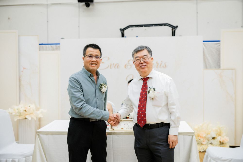 Two men in formal attire shake hands warmly at a wedding reception ceremony.