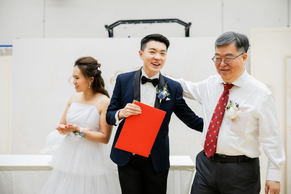 Smiling groom holds red wedding certificate folder alongside bride and elder officiant.