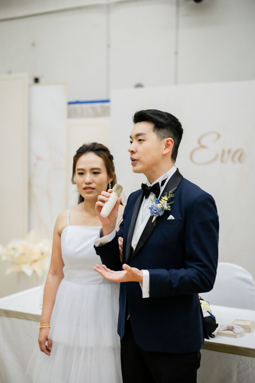 Groom speaks into microphone beside bride Eva during their wedding reception speech.