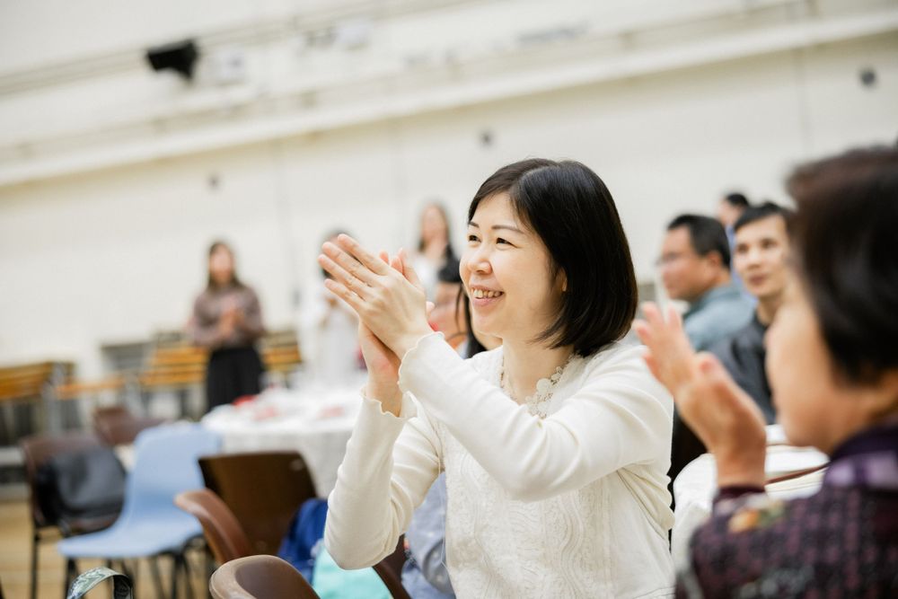 Smiling Asian woman in white sweater clapping joyfully at a wedding reception celebration.