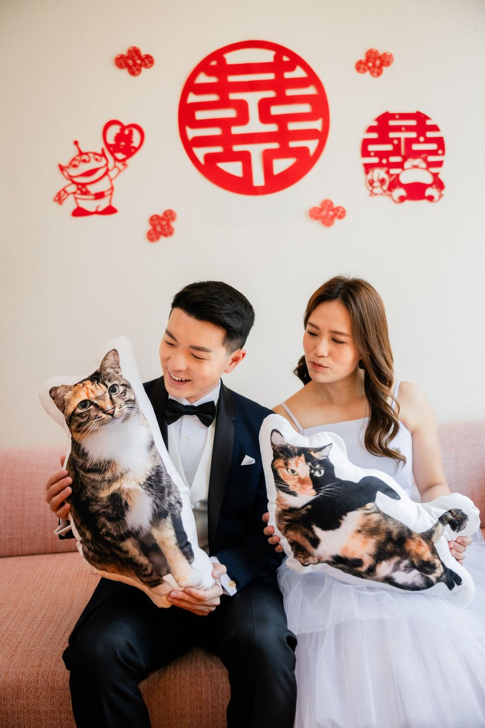 Asian wedding couple sit on pink sofa holding custom cat-shaped pillows beneath red double happiness decorations.
