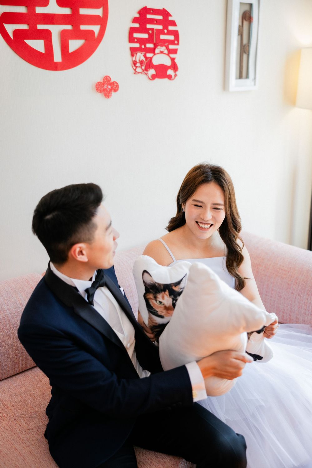 Laughing Asian bride and groom sit on sofa with cat pillow, red Chinese wedding decorations on wall behind.