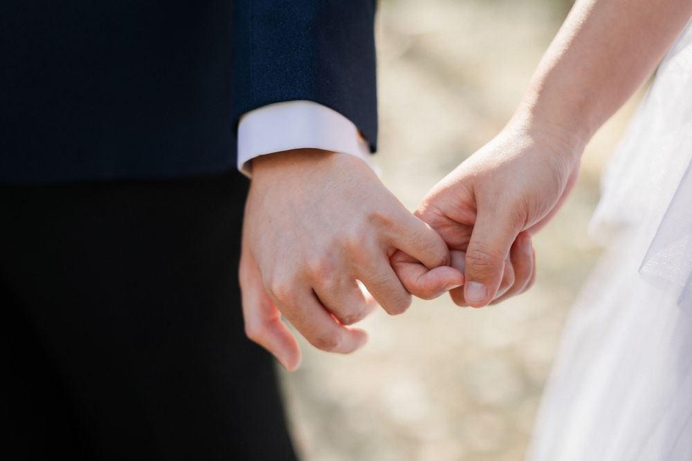 Close-up of bride and groom holding hands outdoors, showing wedding attire details against a soft blurred background.