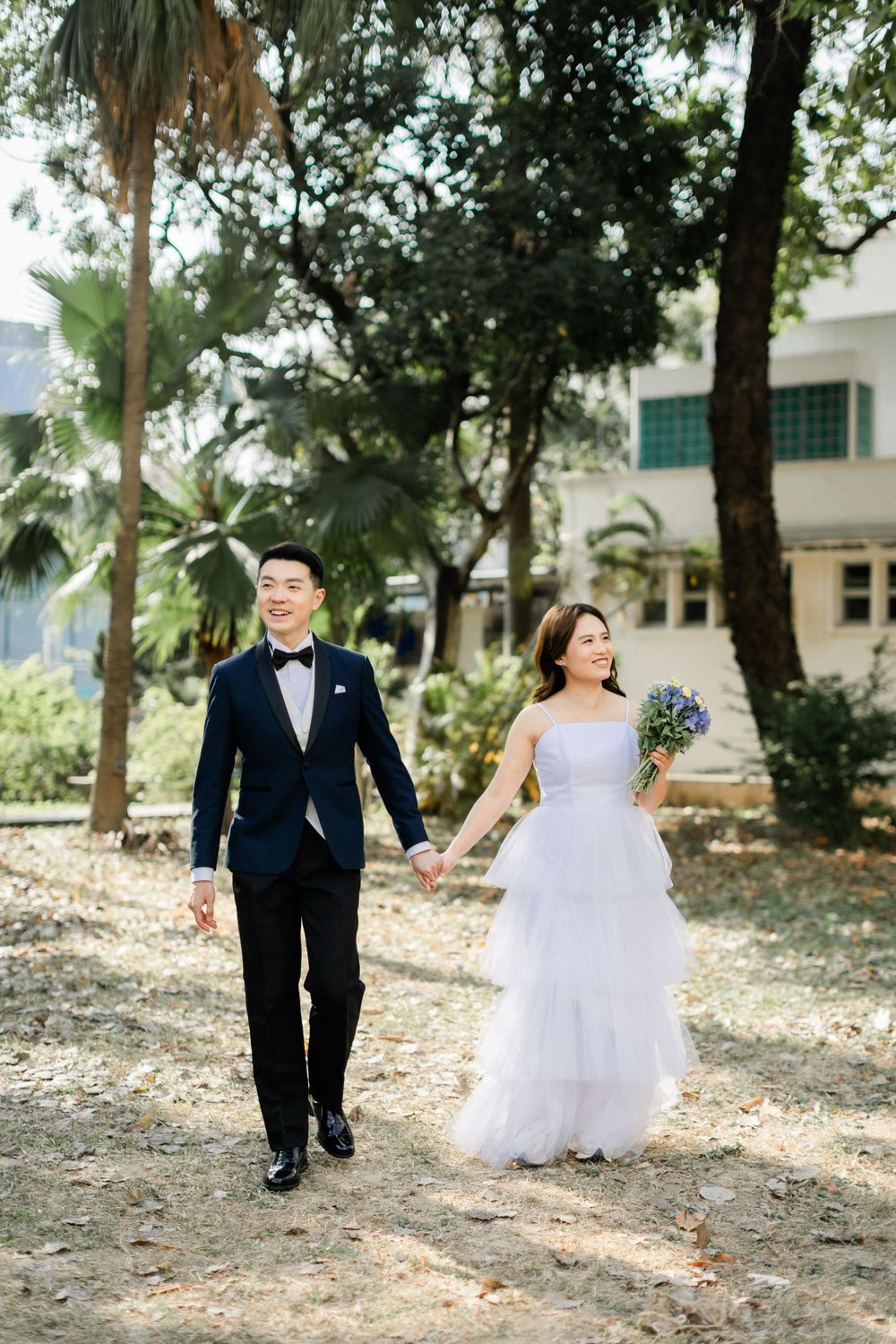 Happy Asian bride and groom walk hand-in-hand through a sunlit park, bride carries a purple and white bouquet.