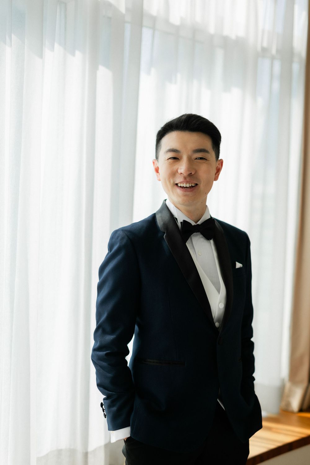Smiling Asian groom in navy tuxedo with bow tie posing confidently by bright window curtains.