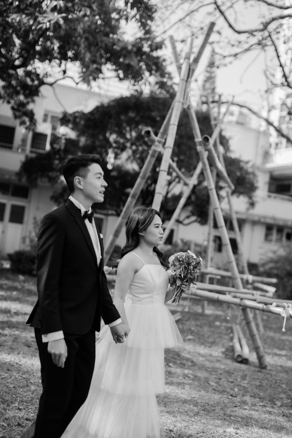 Black and white photo of Asian wedding couple walking hand-in-hand outdoors with trees and building in background.