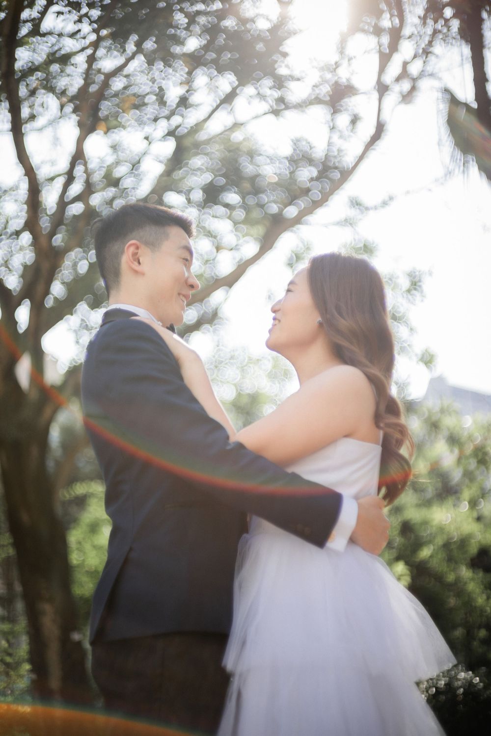 Joyful Asian bride and groom embrace and smile at each other under sunlit trees with dreamy lens flare effect.