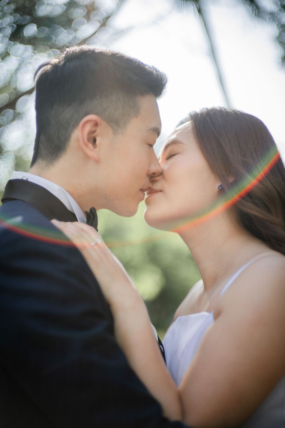Asian bride and groom share a romantic kiss outdoors with soft sunlight and colorful lens flare in background.