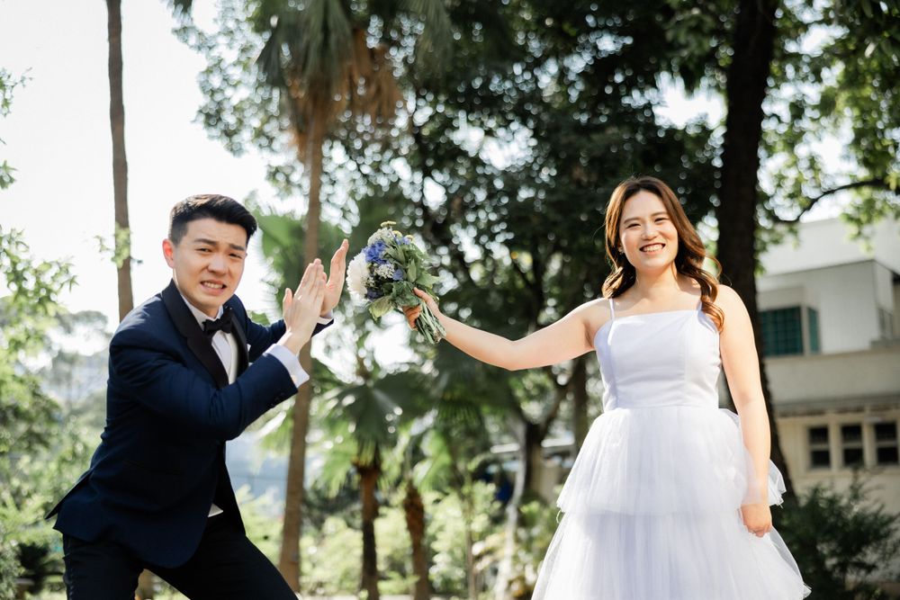 Playful Asian bride and groom high-five outdoors in a sunny park, both smiling joyfully in wedding attire.