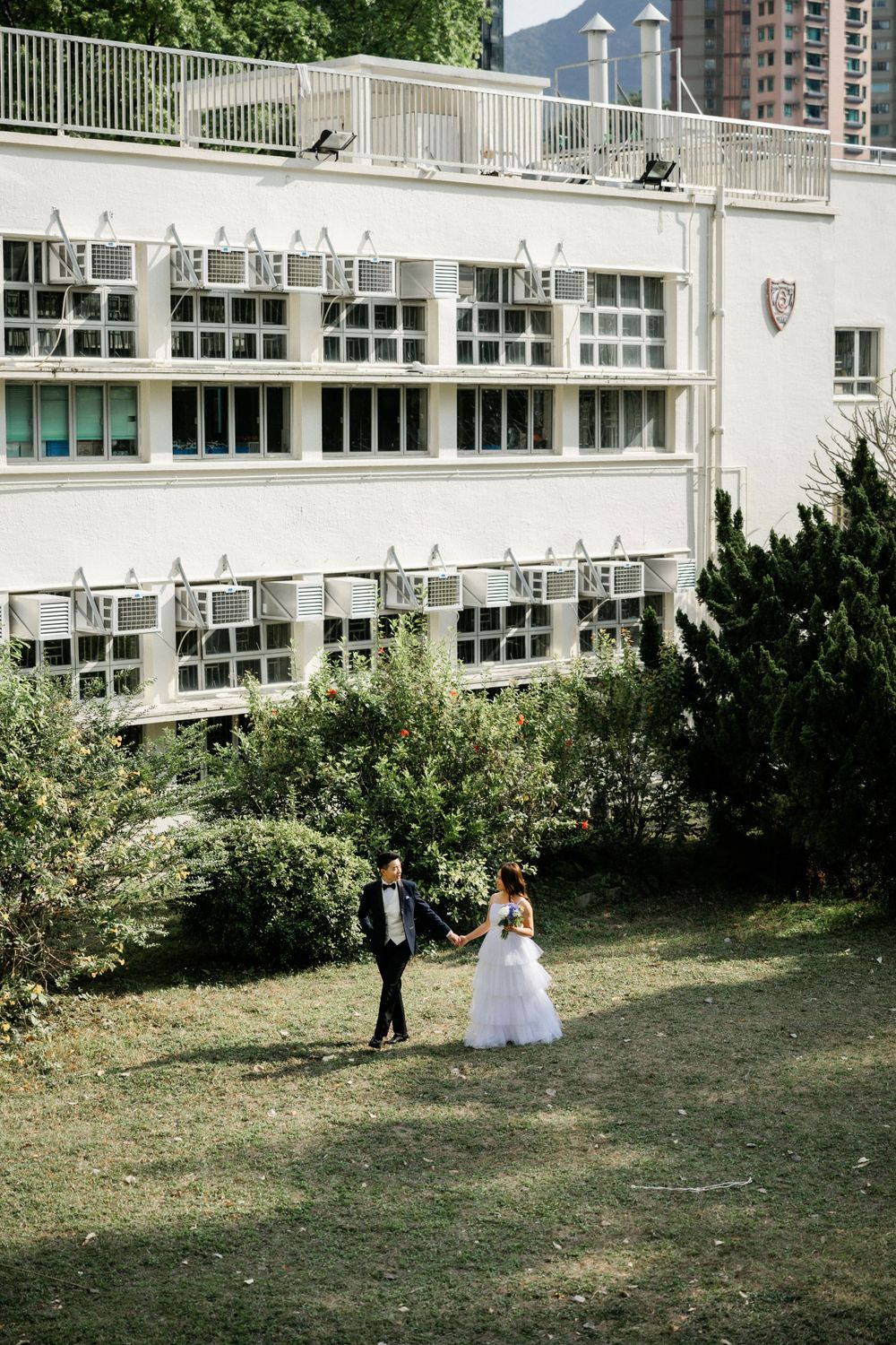 Bride and groom walk hand in hand across green lawn beside white multi-story building with lush trees.
