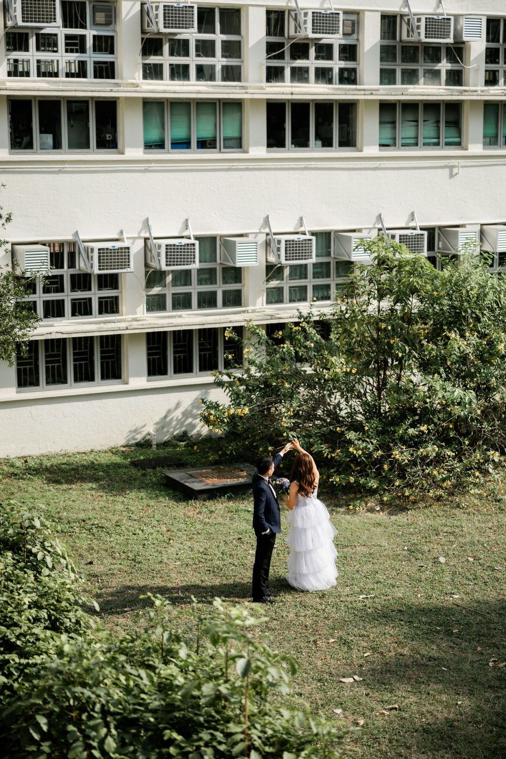Groom twirls bride in white gown on grass lawn in front of building with air conditioning units.