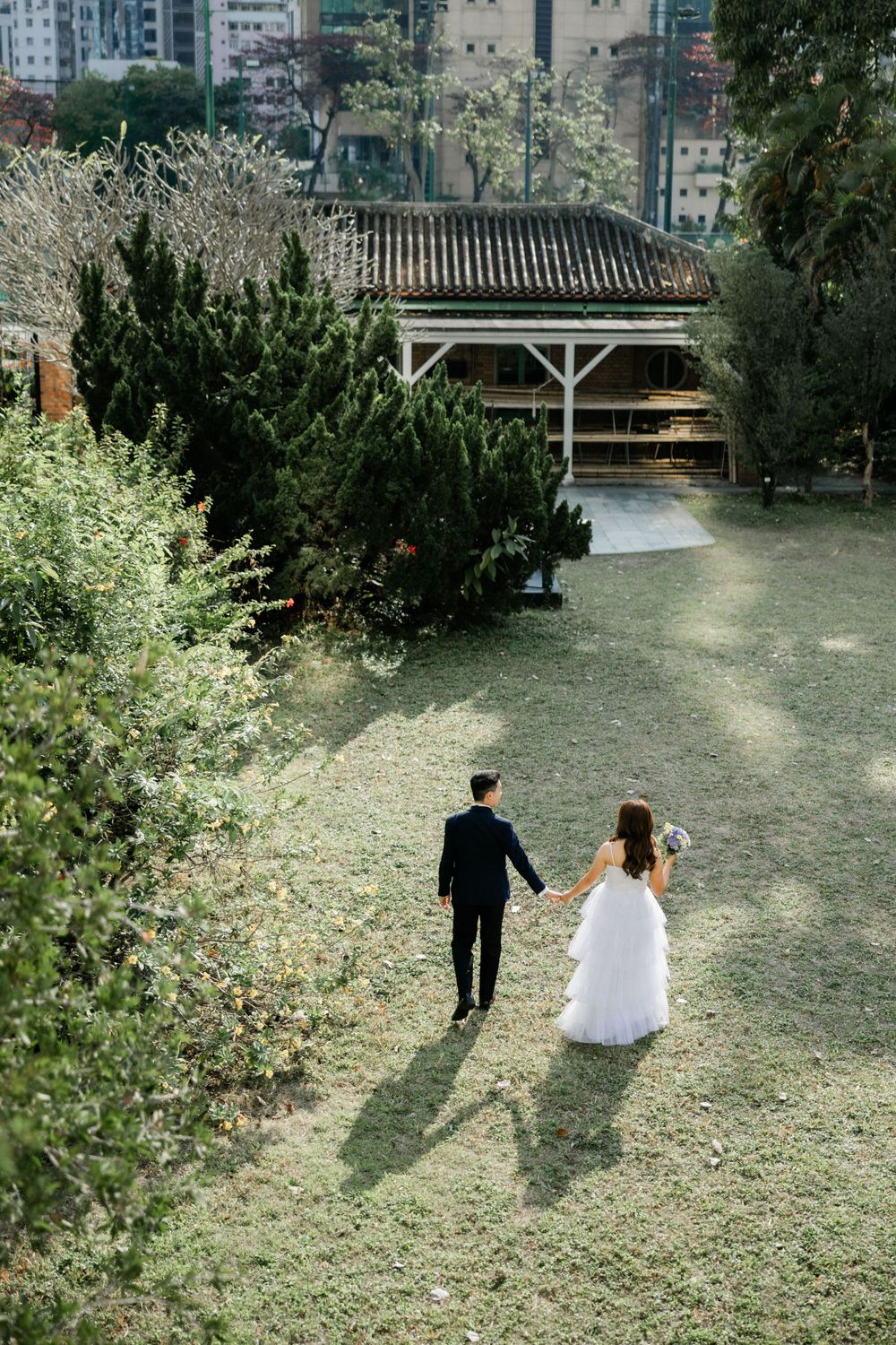 Aerial view of bride and groom holding hands walking toward wooden pavilion surrounded by tall green trees.