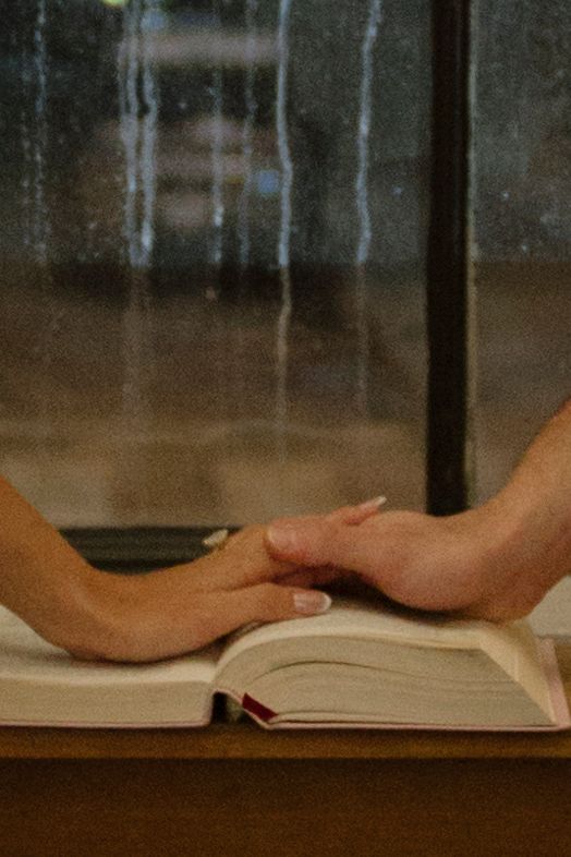 Two hands reach across an open book on a wooden table in an intimate gesture of connection and comfort.