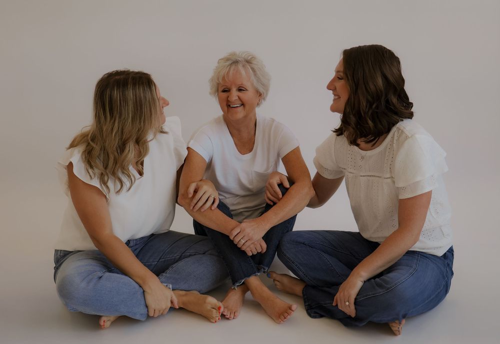 Three women in white shirts and jeans sit cross-legged on floor laughing and sharing joyful moment together.