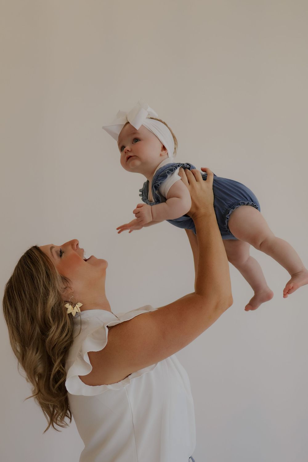 Adult in white top lifts baby wearing blue romper and bow overhead against neutral background.