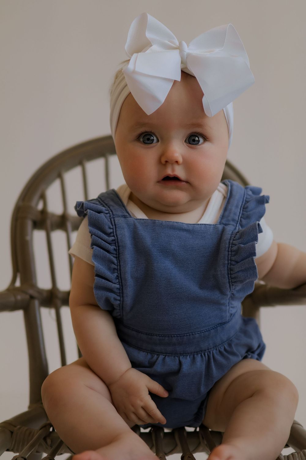 Baby in blue ruffled romper and white bow sits in wooden chair during portrait session.