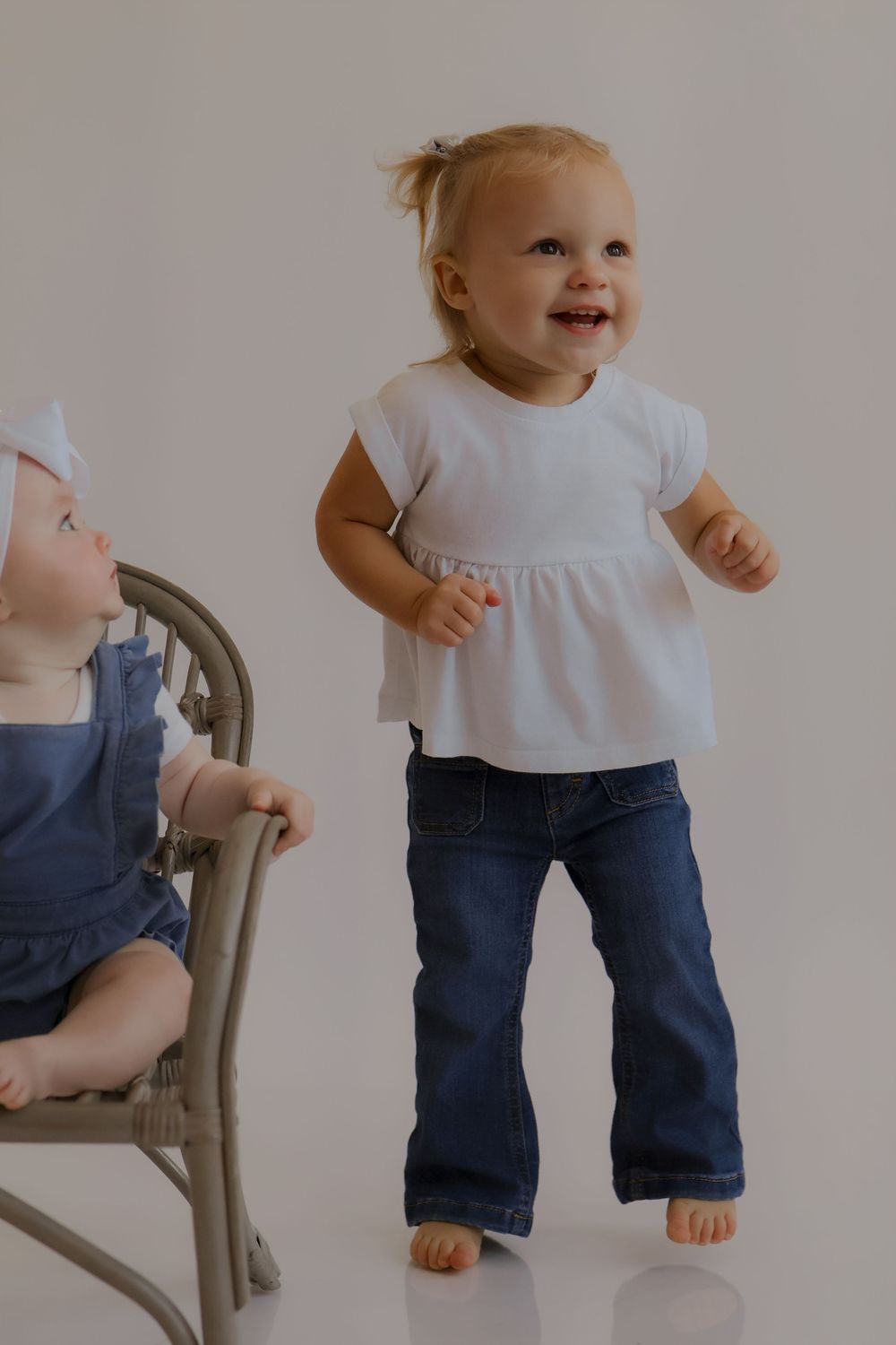 A toddler in jeans and white shirt standing next to a wooden chair against a neutral background.