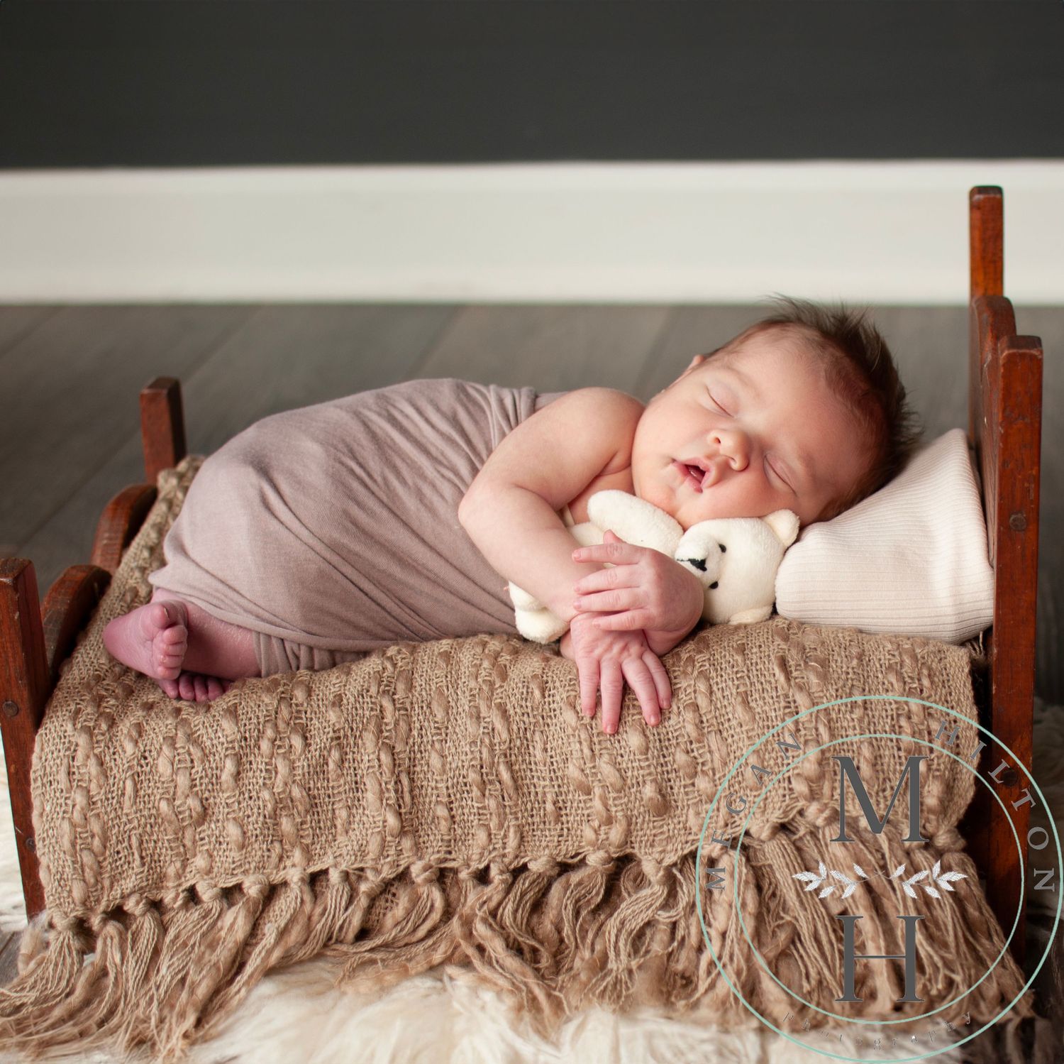 Sleeping newborn wrapped in pale lavender on brown textured blanket with fringe resting on wooden bed.