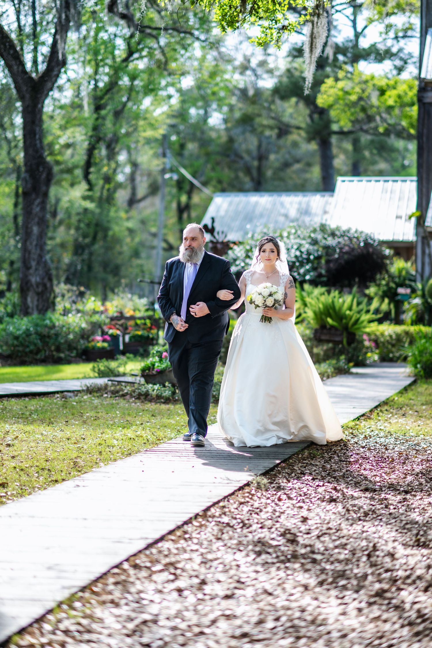 A wedding ceremony taking place on a garden path surrounded by lush greenery on a sunny day.