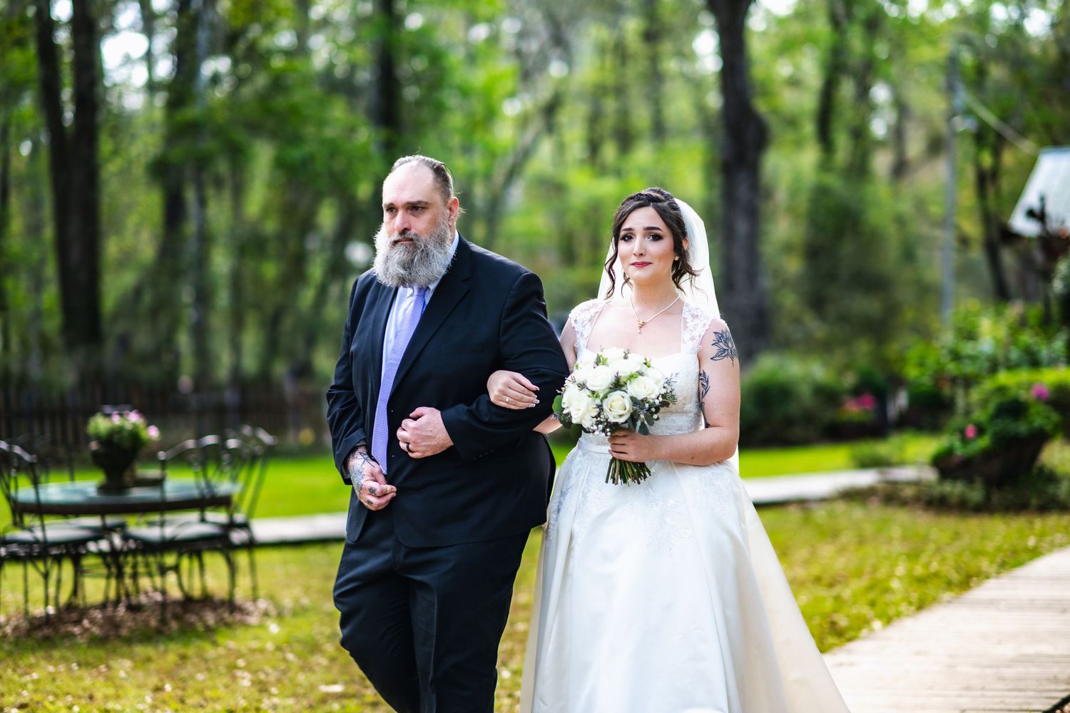 A traditional wedding processional at an outdoor wooded venue with chairs set up among trees in the background.