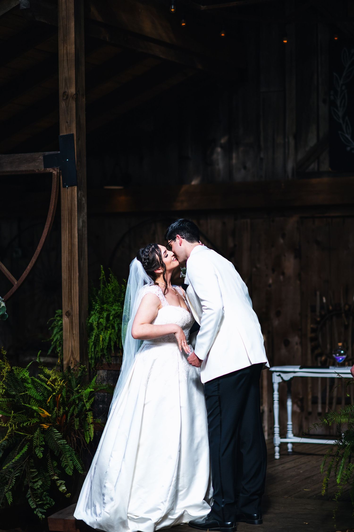 A couple shares a romantic moment in their wedding attire under a rustic wooden shelter surrounded by greenery.