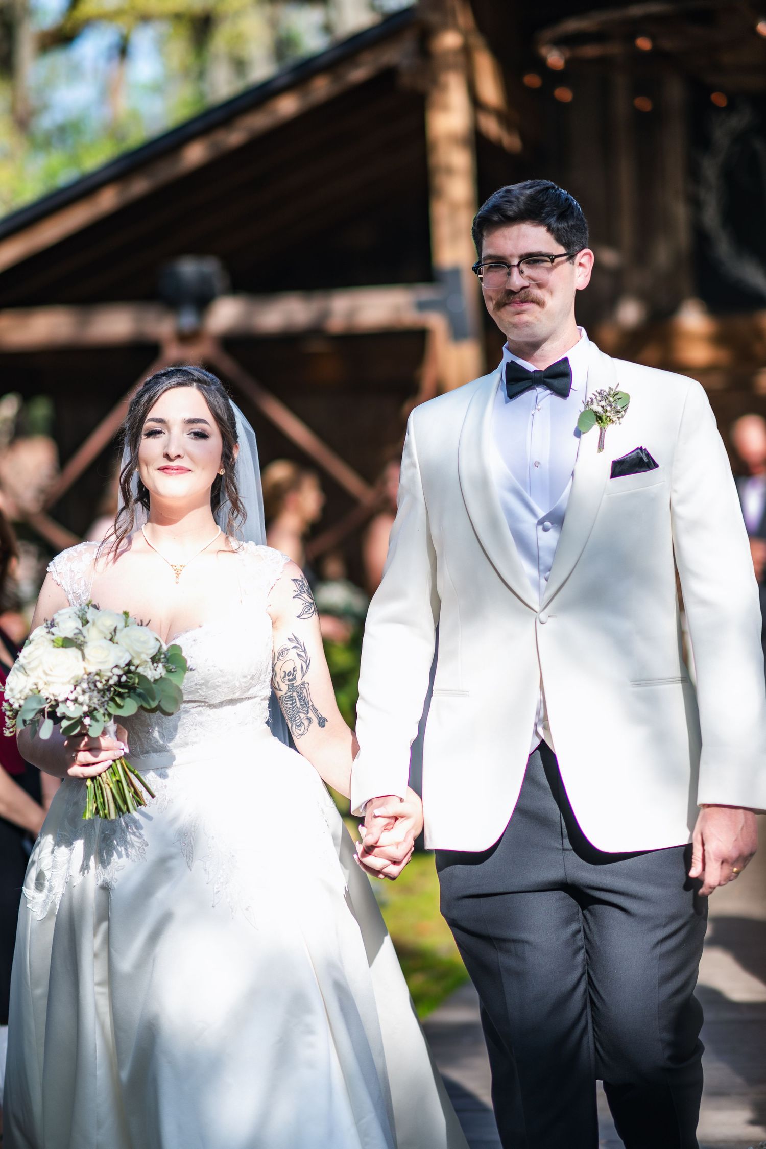 A wedding couple in white attire holds hands outdoors, with the bride wearing a tattoo and holding a floral bouquet.