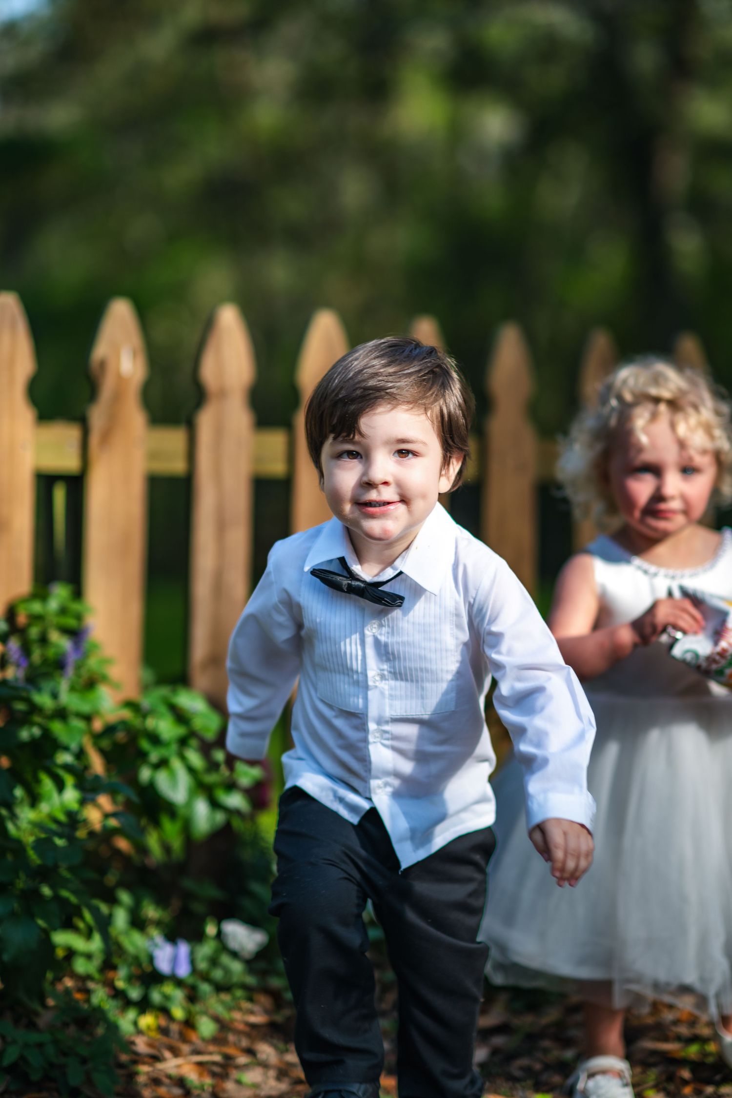 Children dressed in formal attire running and playing in a garden with a wooden fence in the background.