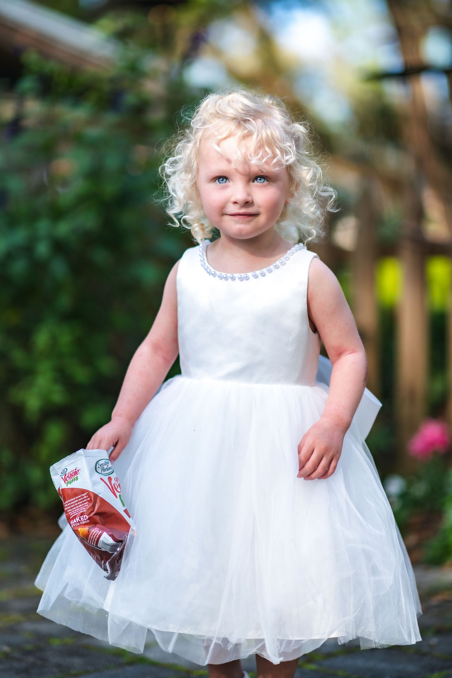 Child wearing a white tulle party dress stands outdoors holding a beverage in a garden setting.