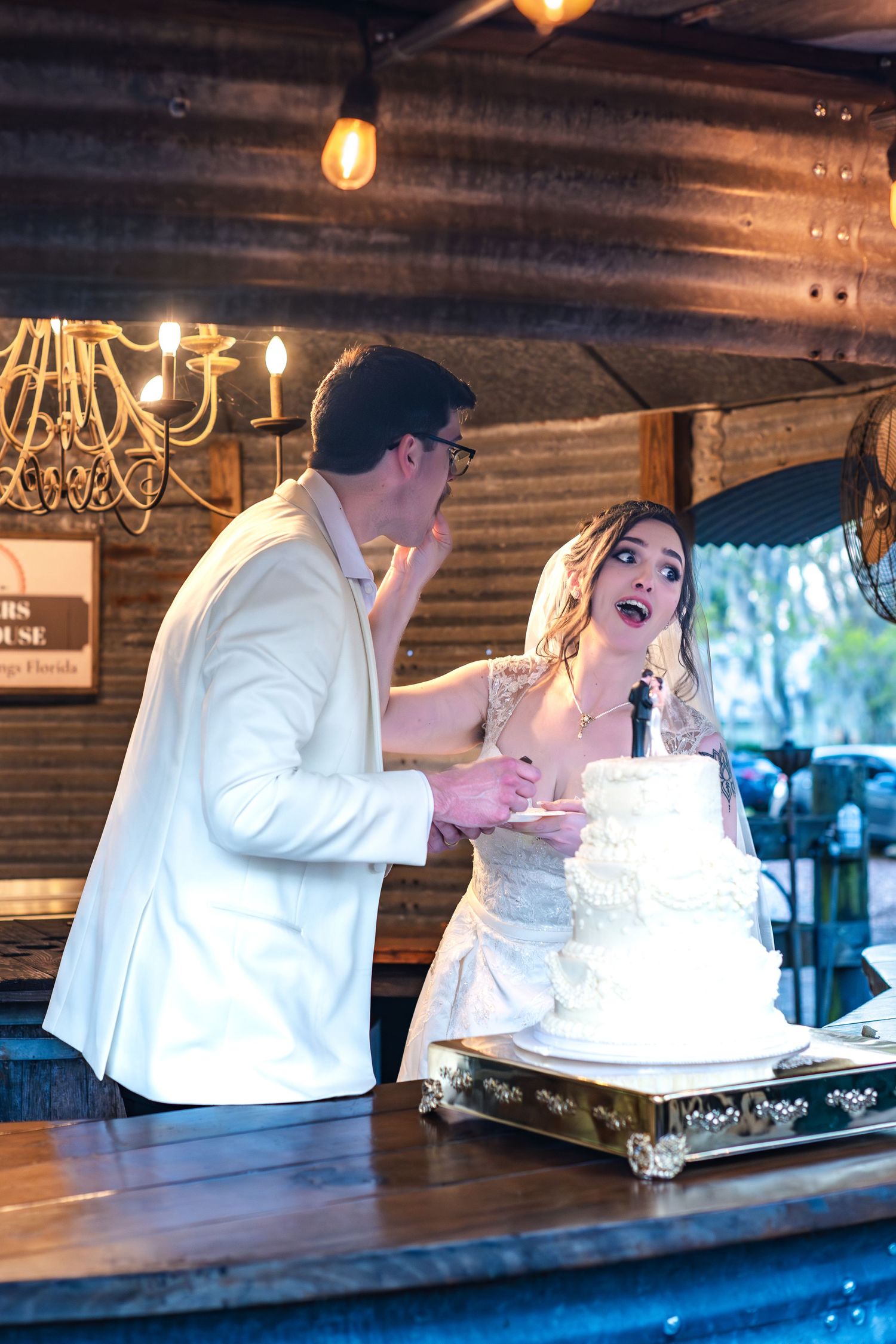 A couple shares a joyful moment while cutting their elegant white wedding cake at a rustic venue with warm lighting.