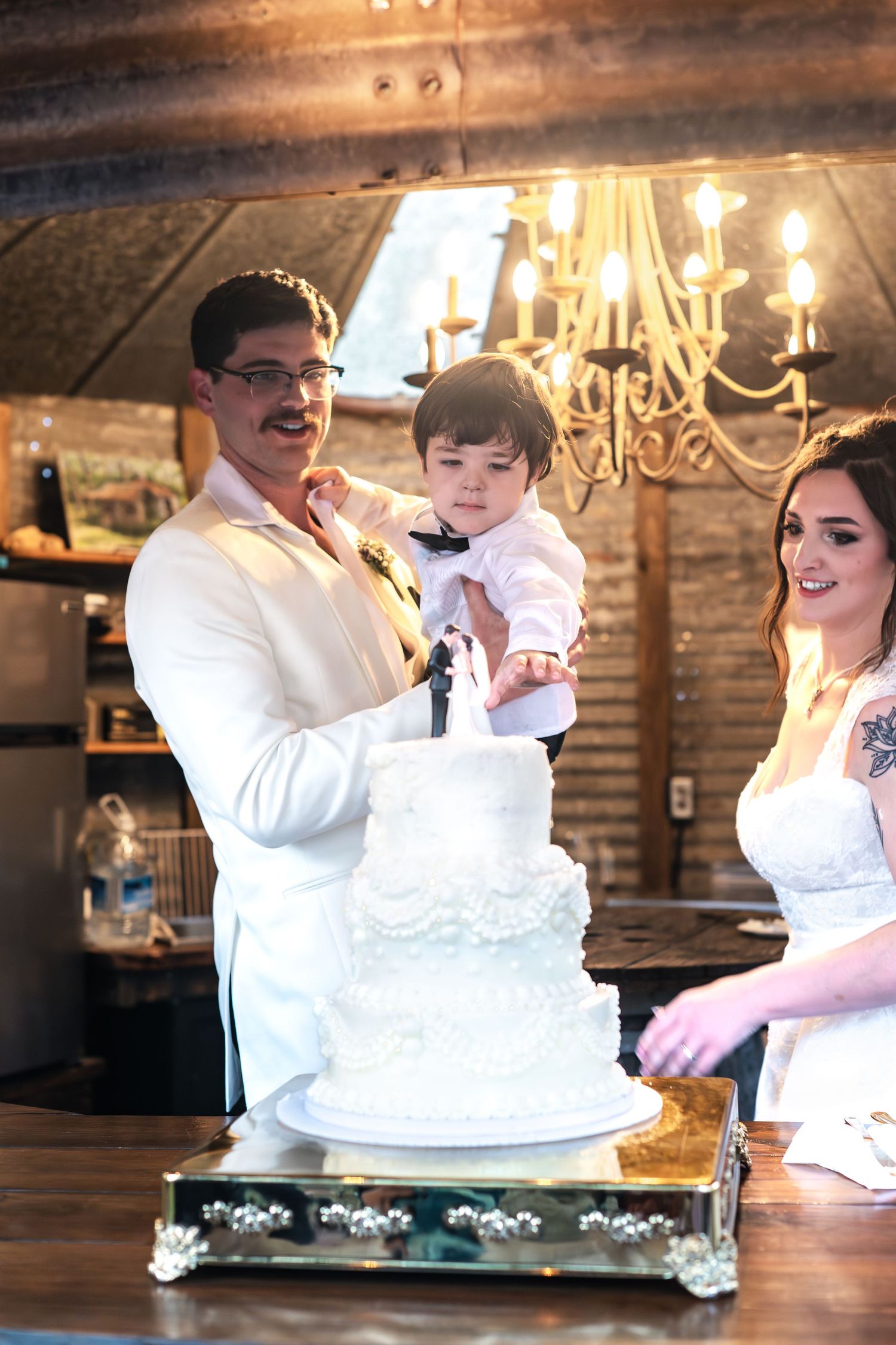 Couple and small child celebrate at a wedding reception with an elegant white tiered cake under a chandelier.