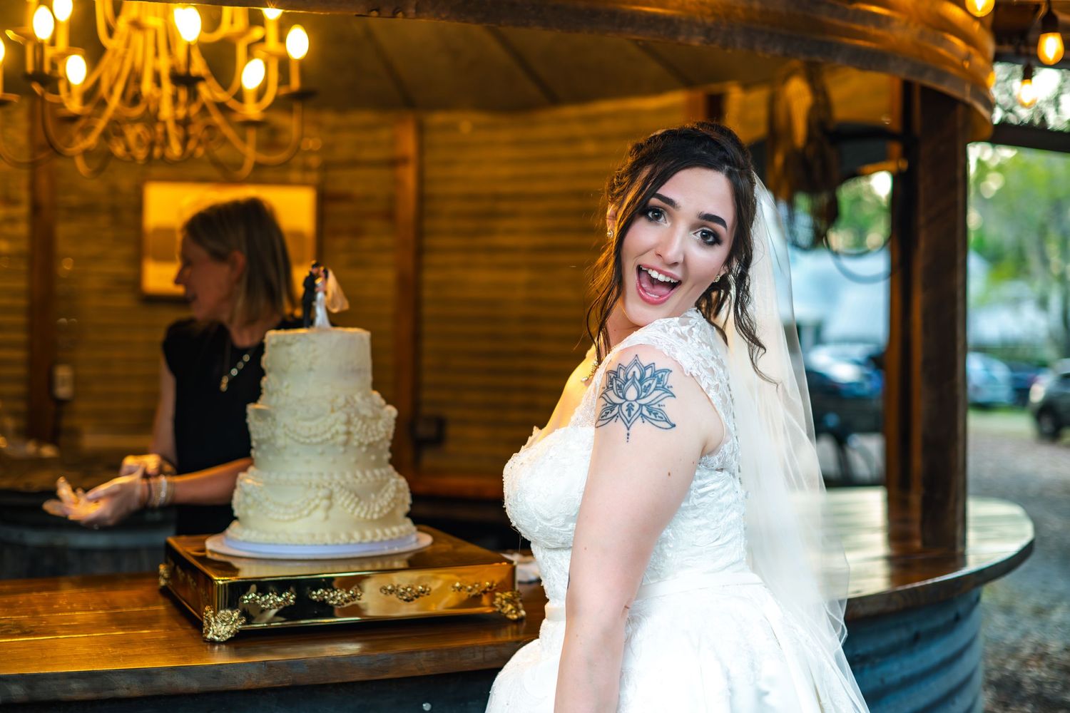 A bride in a white wedding dress with tattoos poses beside a rustic white wedding cake at a wooden venue.